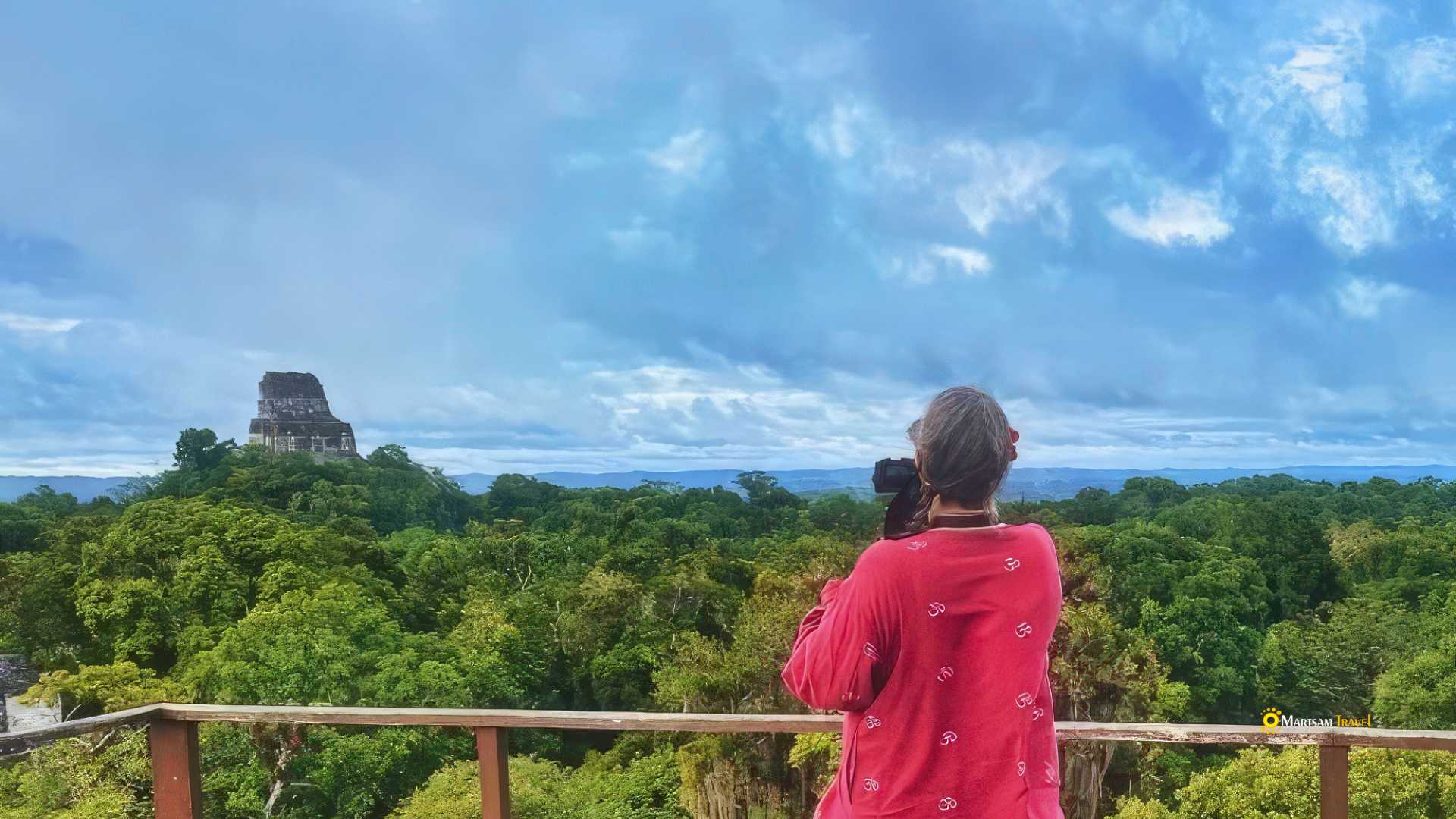 Lady atop a Mayan pyramid at Tikal, gazing over the vast tropical forest canopy, with ancient temples rising majestically amidst the tranquility.