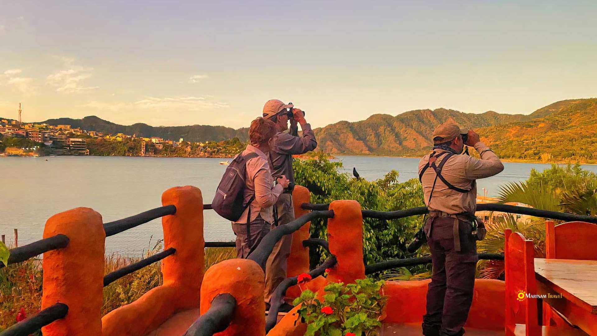 Couple using binoculars for birdwatching along the serene shores of Lake Atitlan, with the lake's clear waters and surrounding mountains in the background.