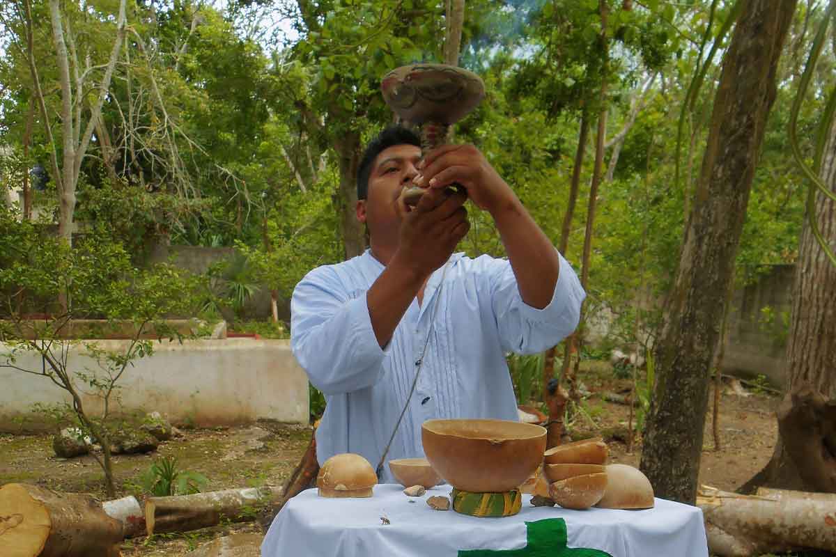 An image of a Mayan shaman dressed in traditional ceremonial attire, holding a wooden smoking cup in both hands. The shaman is surrounded by a mystical atmosphere, performing a Maya Ceremony in a natural outdoor setting