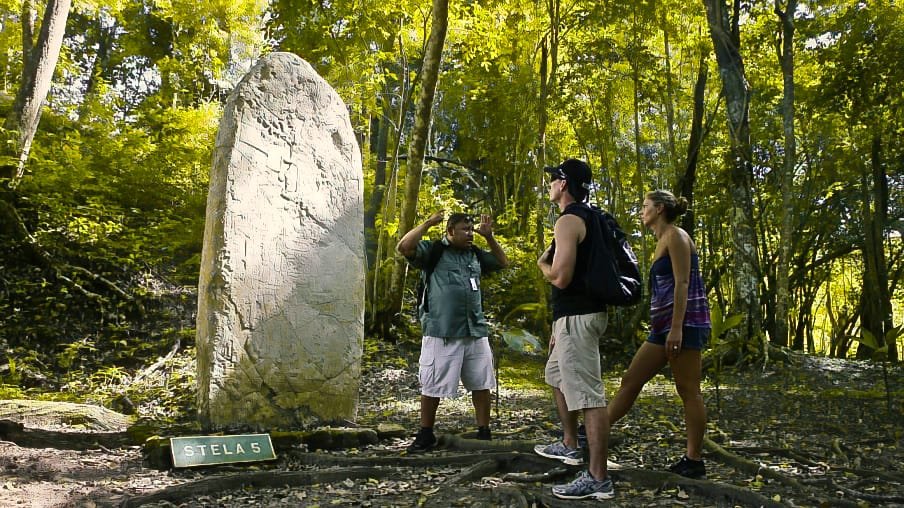 A young couple of travelers listening to a local guide explain Maya history beside an ancient stela in a lush tropical forest, highlighting the cultural depth of vacation packages in Belize.