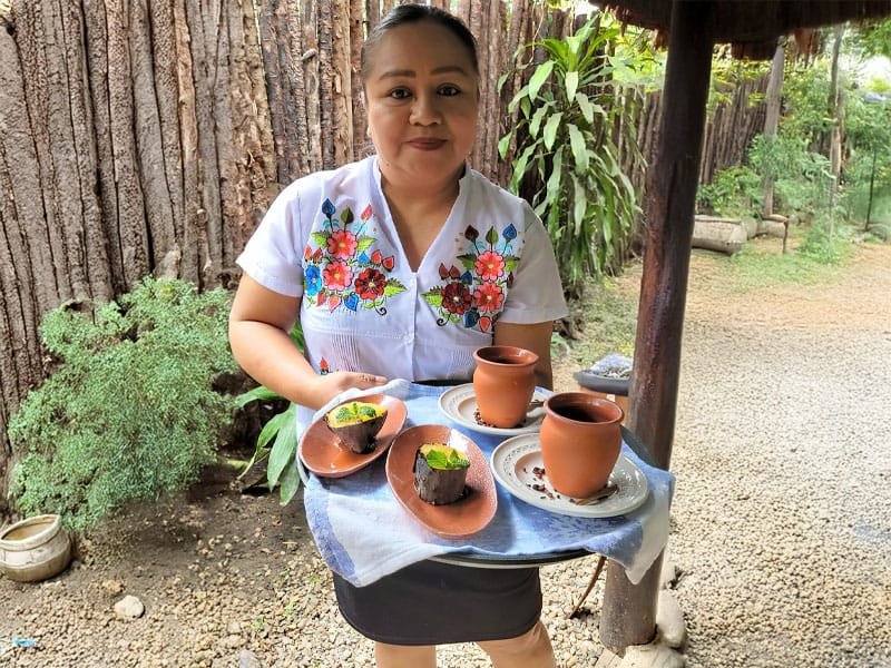 Belizean woman offering a traditional delicacy on a tray, wearing a floral-embroidered blouse and smiling warmly against a local backdrop.