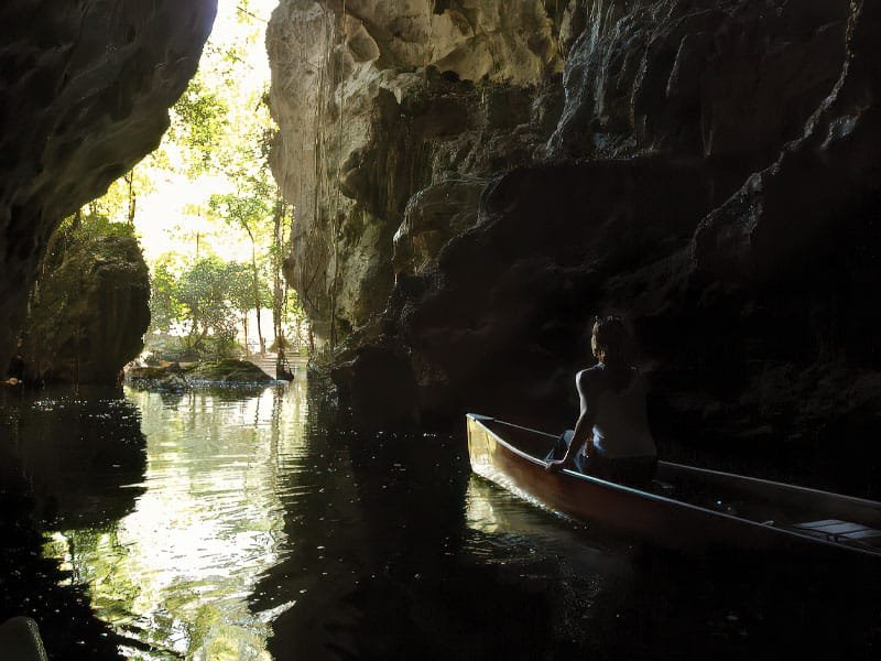 Woman traveling in a canoe inside a sacred cave in Belize, surrounded by mystical rock formations and dim natural light.