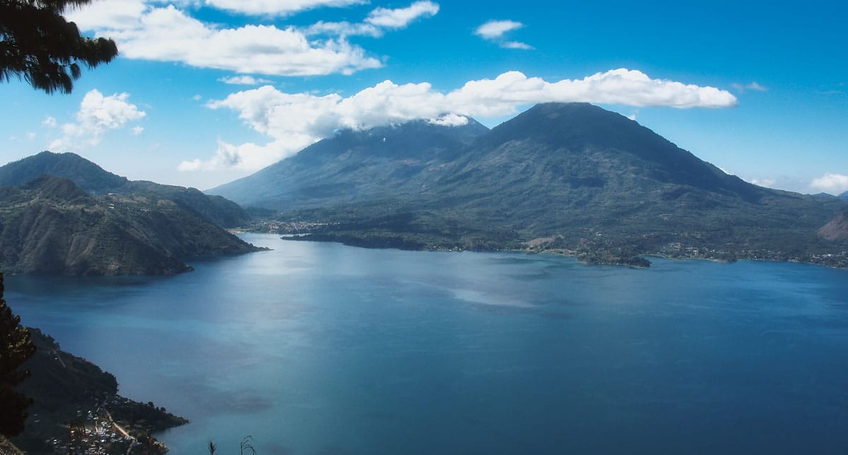 Scenic view of Lake Atitlán with calm blue waters, surrounded by lush hills and majestic volcanoes under a clear sky.