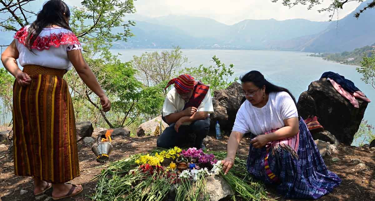 Mayan people, including two women and a shaman, perform a traditional Maya ceremony by Lake Atitlan, dressed in colorful attire.