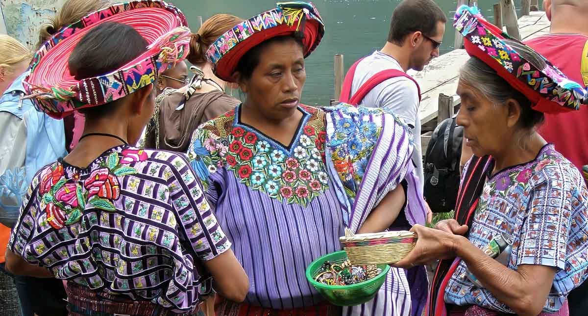 Three Mayan women in colorful traditional attire standing by Lake Atitlan, showcasing rich cultural heritage and vibrant local fashion.