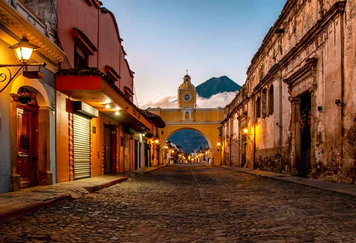 View of Calle del Arco in Antigua Guatemala at dusk, with the Agua Volcano in the background and the first streetlights glowing—capturing the charm of one of the best private tour company destinations in Guatemala from the US.