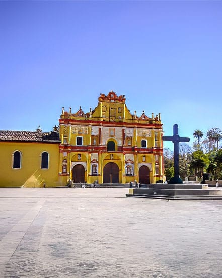 Yellow Baroque church with three arches in a spacious plaza, a tall blue cross sculpture to the right, with palm trees and a clear blue sky in the background.