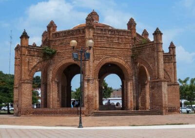 Brick arch gateway with multiple arches in a spacious square, a vintage lamppost in the foreground and a bright blue sky above.
