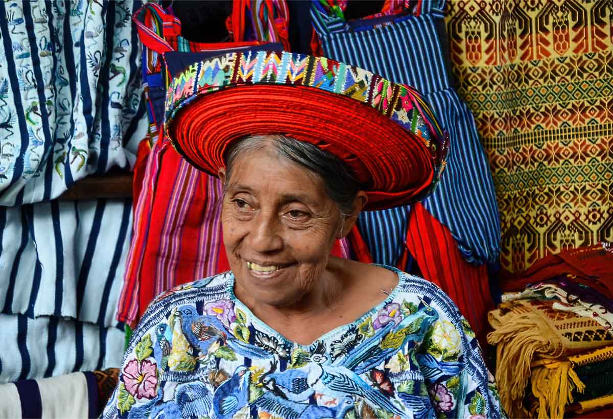 Maya-tzutujil | martsam travel Tz’utujil Maya woman wearing traditional clothing beside woven textiles in Guatemala.