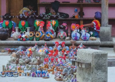 Colorful ceramic animal figurines arranged on a stone ledge and ground at a market stall, with a vendor seated near a pink wall in the background.