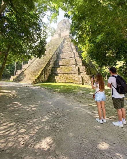 Two travelers photographing a Maya temple beneath the trees at Tikal.