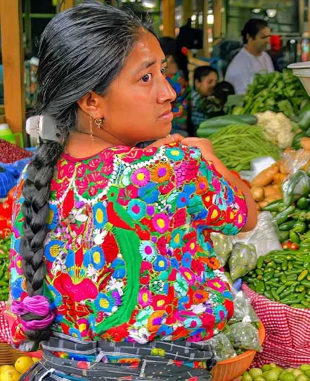 Woman wearing a colorful embroidered blouse beside a vegetable stall in a Guatemalan market.