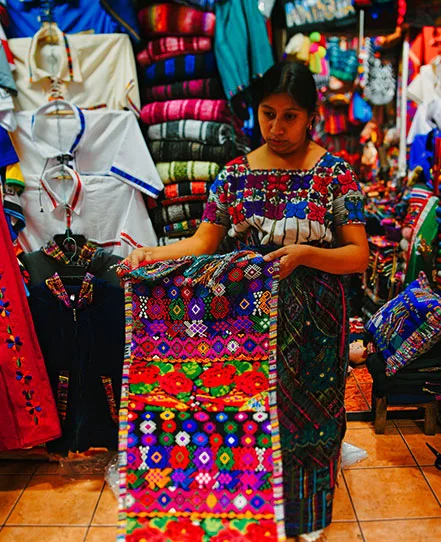 Woman displaying a brightly patterned textile inside a shop filled with woven clothing in Guatemala.