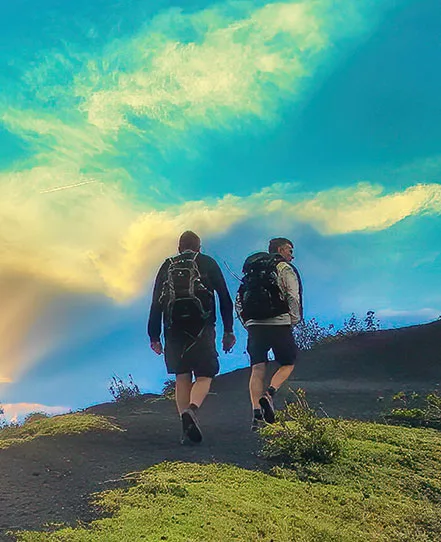 Two hikers with backpacks walking uphill beneath a colorful sky in Guatemala.