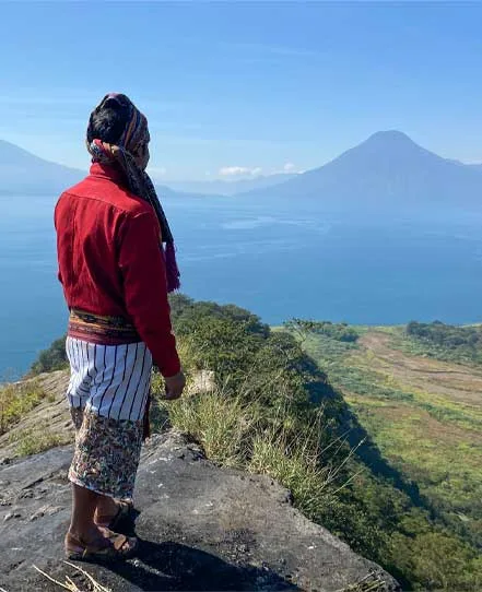 Maya man in a red blouse standing at a rocky viewpoint above Lake Atitlán and its surrounding volcanoes.