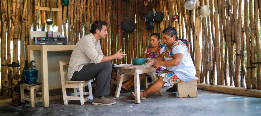 Two Maya women preparing food at a wooden table while a visitor joins them in their kitchen.
