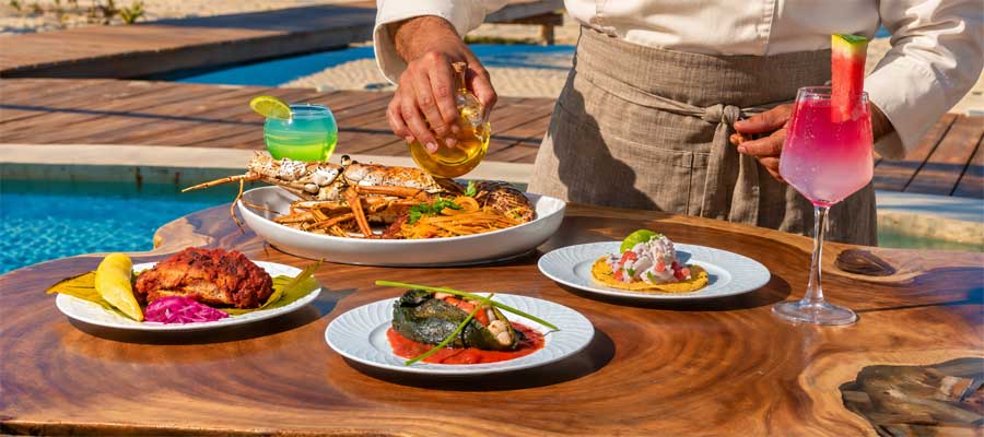 Chef in an ivory jacket finishing a lobster dish beside a pool, with plated specialties and cocktails.