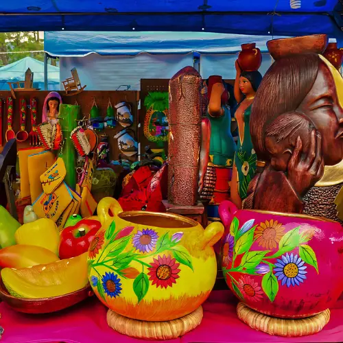 Incentive-travel-el-salvador-artisan-market | martsam travel Colorful painted ceramic pots with floral designs displayed at a market stall, with carved wooden figures and crafts in the background.